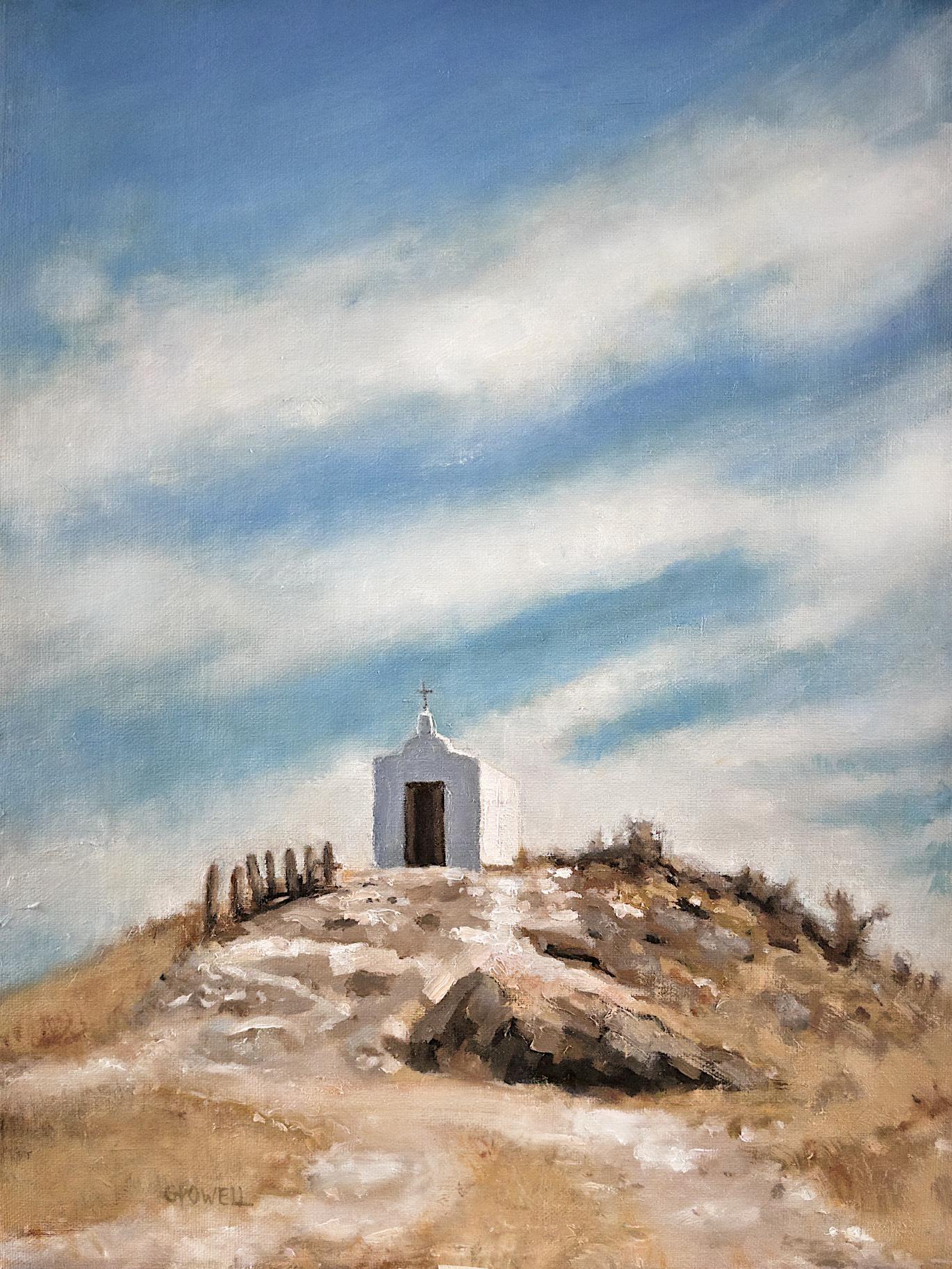 A small white chapel sits on top of a rocky, dry hill. The sky is expansive and dramatic with streaky white clouds. The colors are mostly blue, brown and neutral yellow-oranges.