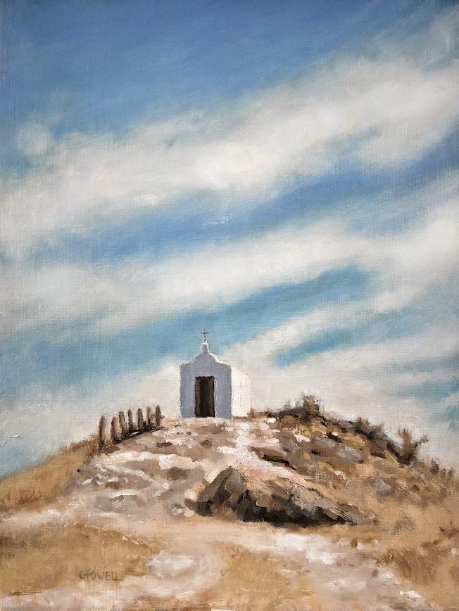 A small white chapel sits on top of a rocky, dry hill. The sky is expansive and dramatic with streaky white clouds. The colors are mostly blue, brown and neutral yellow-oranges.