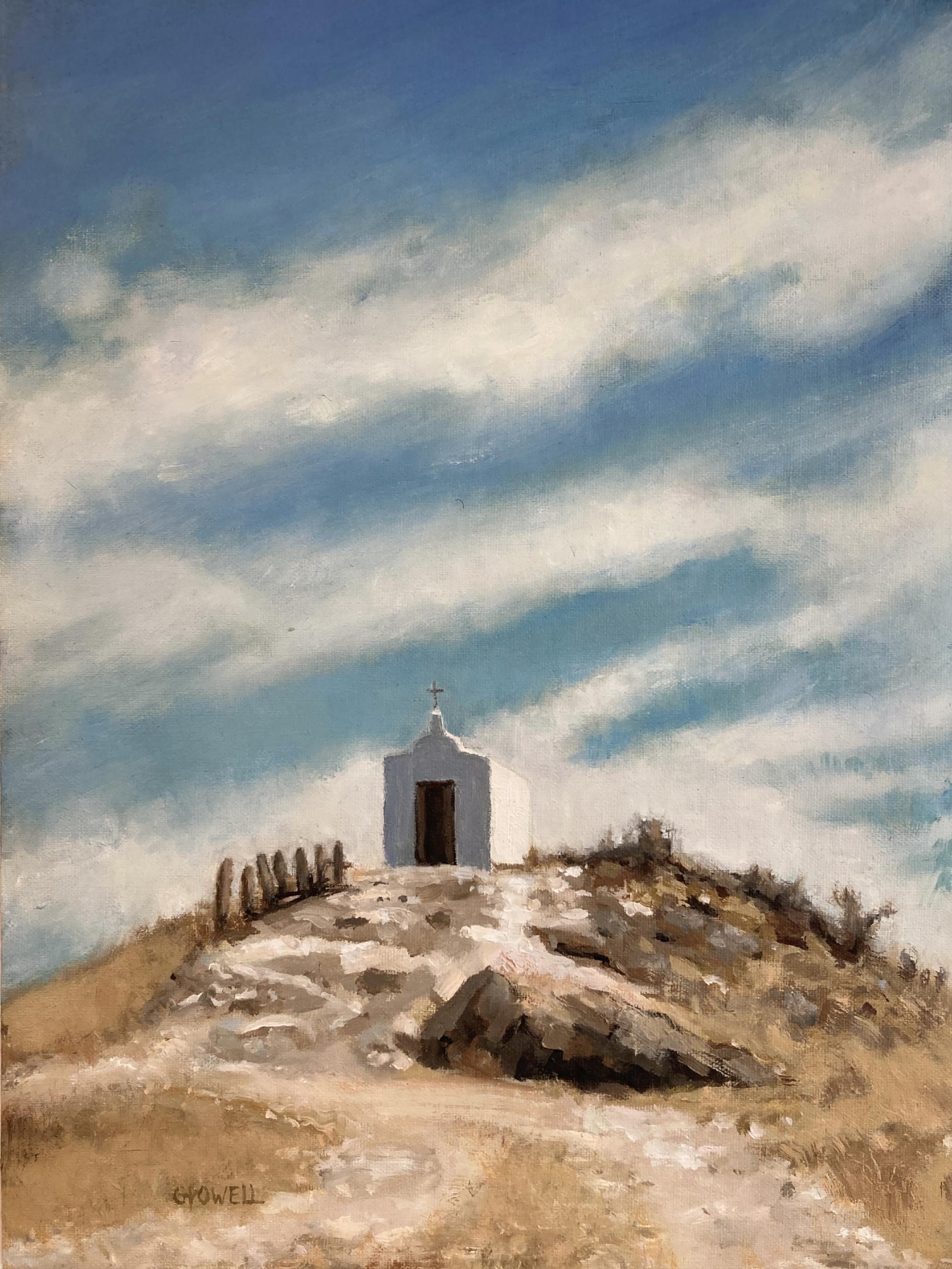 A small white chapel sits on top of a rocky, dry hill. The sky is expansive and dramatic with streaky white clouds. The colors are mostly blue, brown and neutral yellow-oranges.