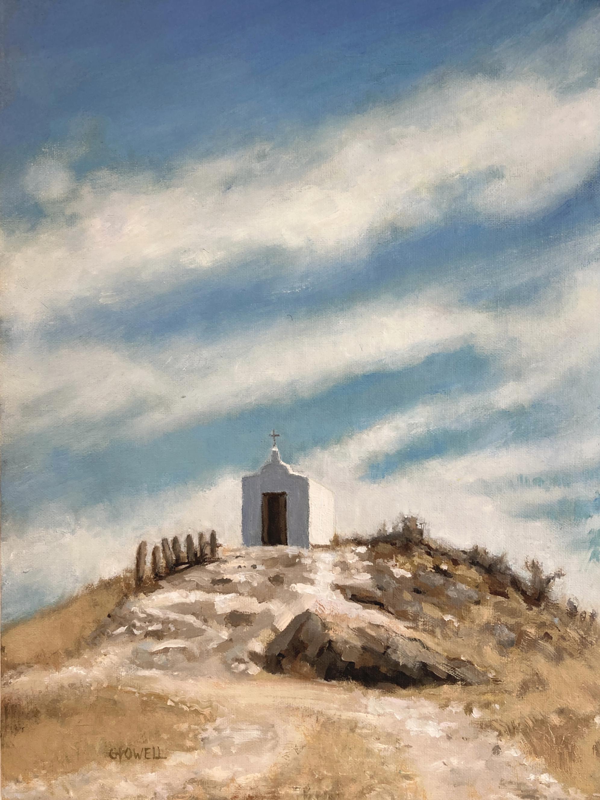A small white chapel sits on top of a rocky, dry hill. The sky is expansive and dramatic with streaky white clouds. The colors are mostly blue, brown and neutral yellow-oranges.