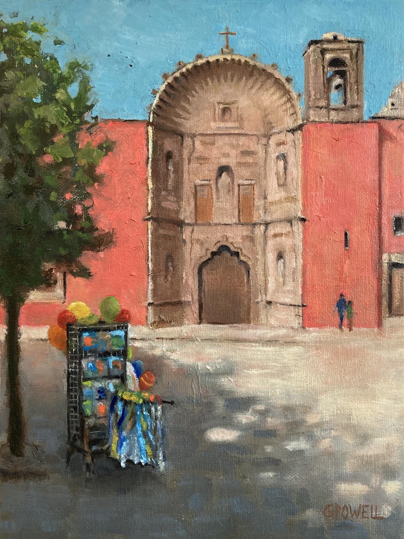 A seller's kiosk sits idly, full of colorful toys in the shade of a tree, while the facade of a temple is visible in the background below a clear blue sky. Its walls are a vibrant orange-red.
